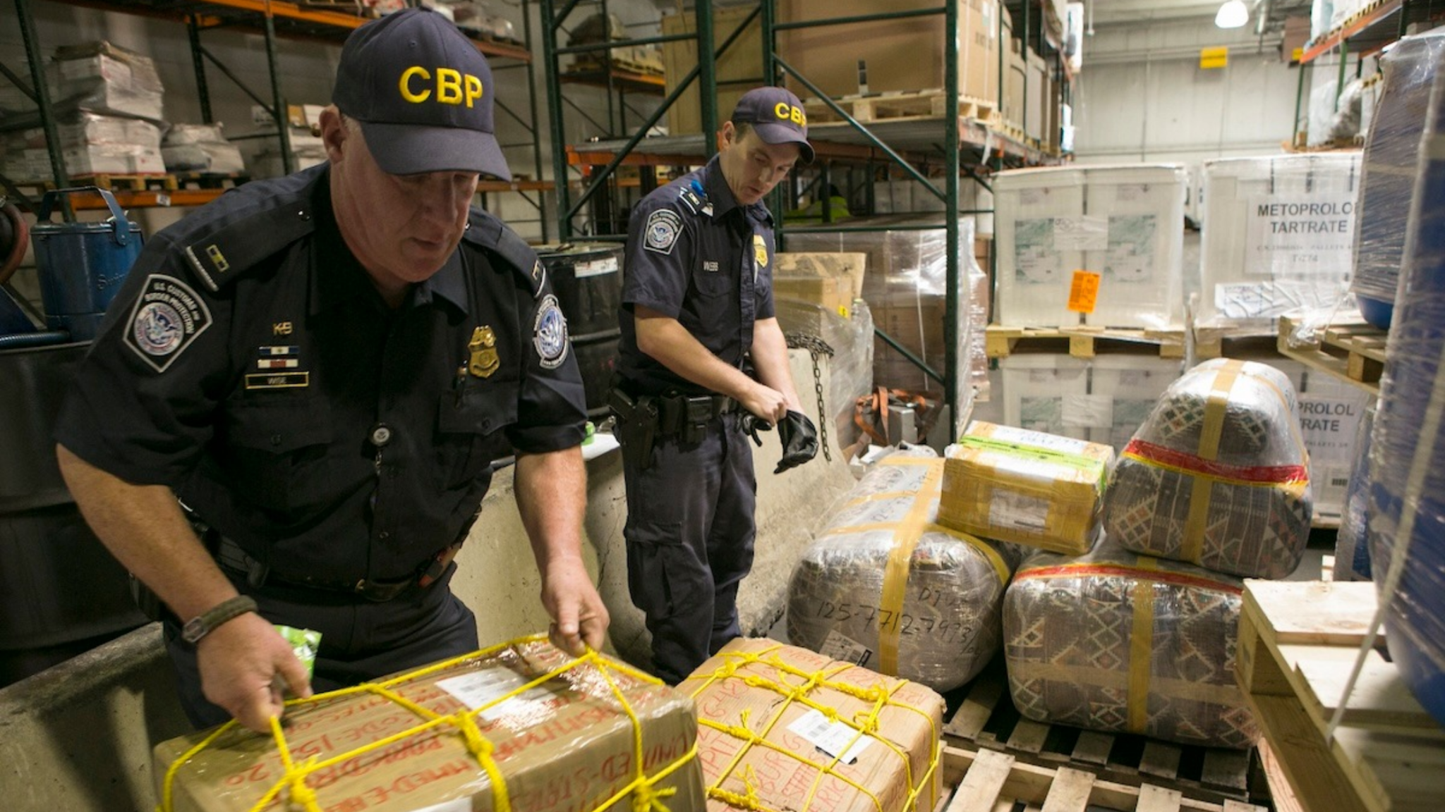 In a large warehouse, two U.S. Customs and Border Protection officers in uniform stand over several packages wrapped in yellow mesh as they inspect a shipment of drugs and chemicals.